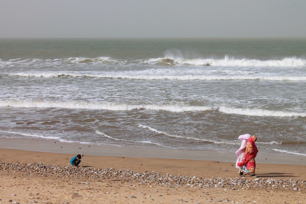 two women in saharan wraps walking along coastline while little boy plays in sand